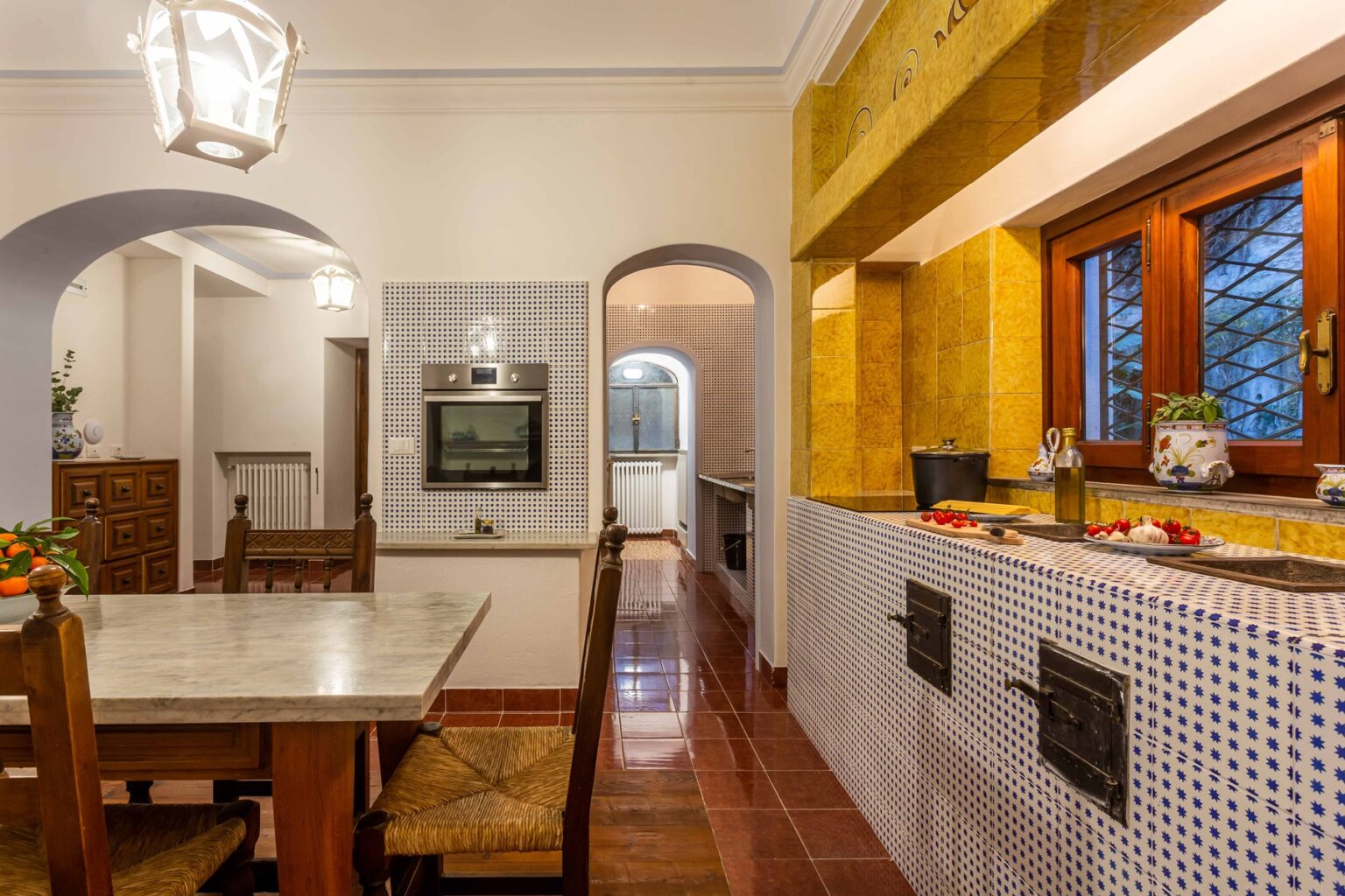 Modern kitchen and dining area featuring a marble table, wooden chairs, and warm lighting. The kitchen showcases yellow tile accents and a tiled backsplash, with an archway leading to additional spaces. Perfect for home decor inspiration. | Only on SopranoVillas
