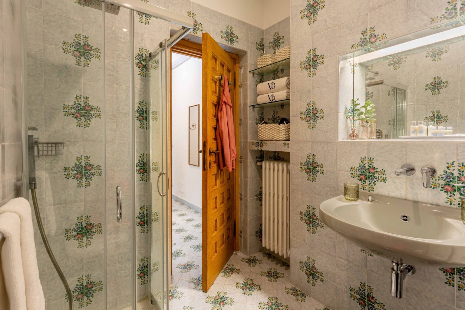 Modern bathroom interior featuring a glass shower, a sleek sink, and decorative tiled walls. The wooden door is slightly open, revealing a neatly organized space with towels and bathroom essentials. Natural light illuminates the room, enhancing its inviting atmosphere. | Only on SopranoVillas