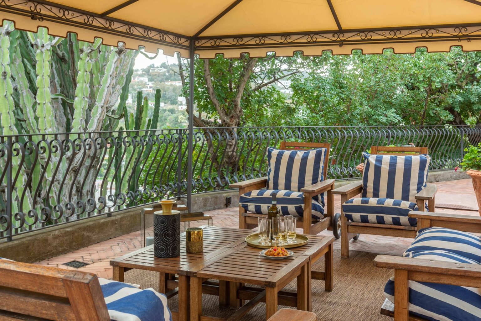 Cozy outdoor seating area with striped cushions, a wooden table set with drinks and snacks, surrounded by lush greenery and a decorative iron railing. Ideal for relaxation and entertaining guests. | Only on SopranoVillas