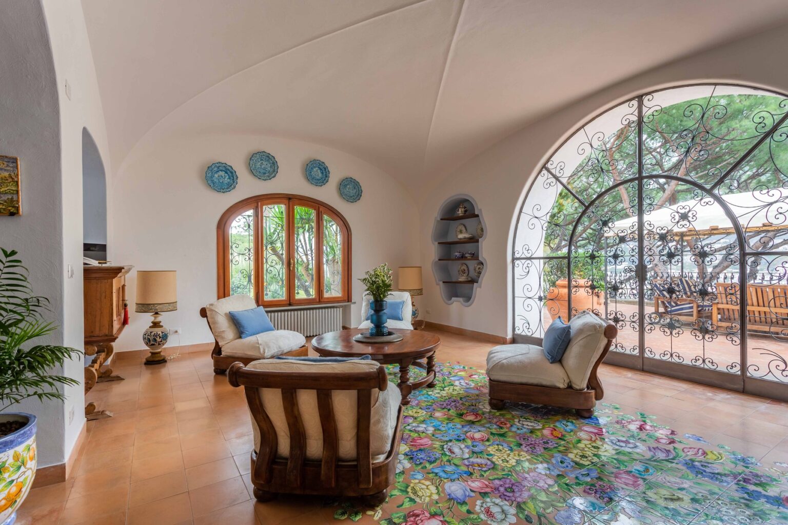 Bright and airy living room featuring comfortable seating, a colorful area rug, and large arched windows that offer natural light and views of the outdoors. Decor includes decorative plates on the walls and indoor plants, creating a welcoming atmosphere. | Only on SopranoVillas