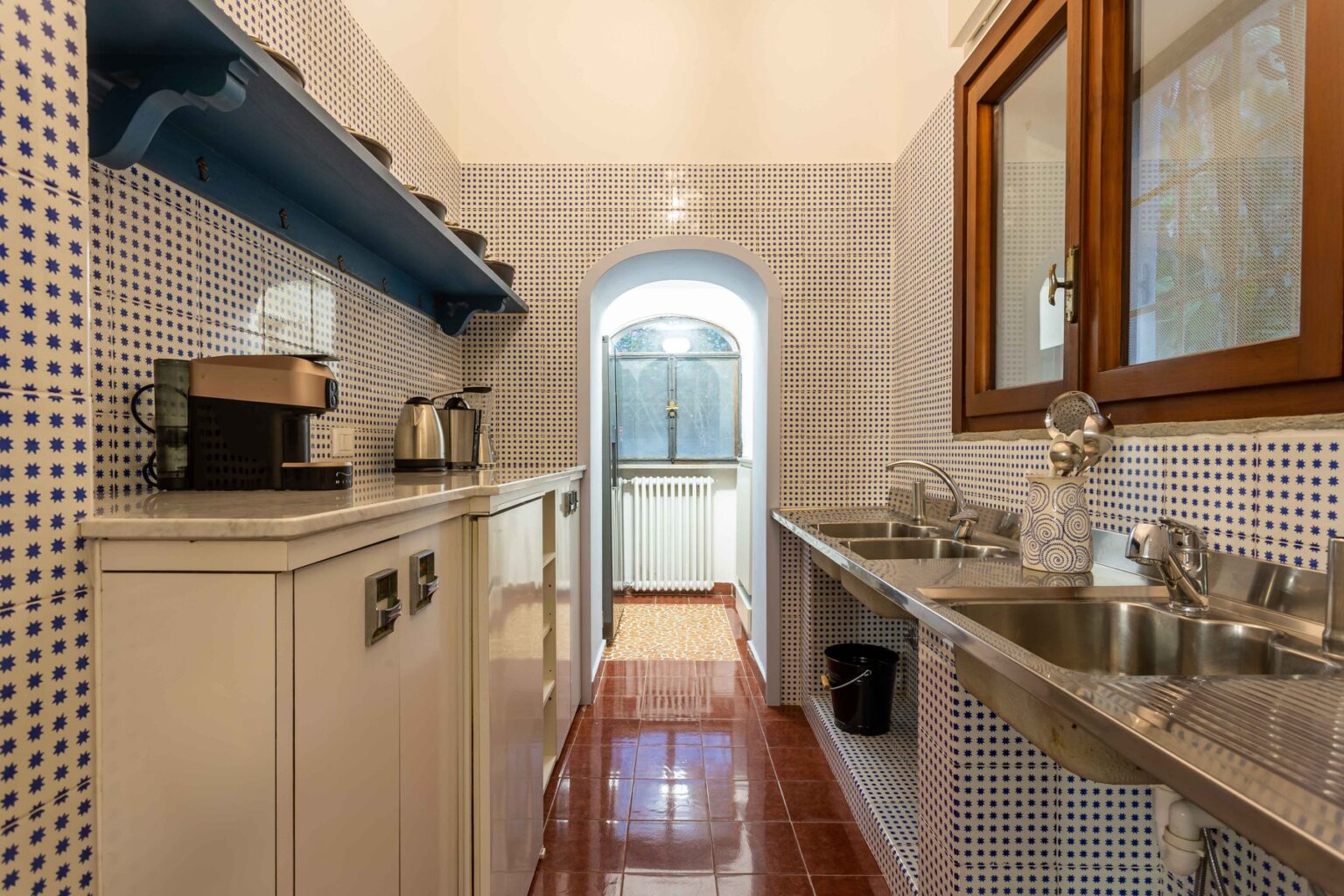 Narrow kitchen with tiled walls, featuring a coffee maker, sink, and wooden cabinets, leading to a doorway at the end. | Only on SopranoVillas