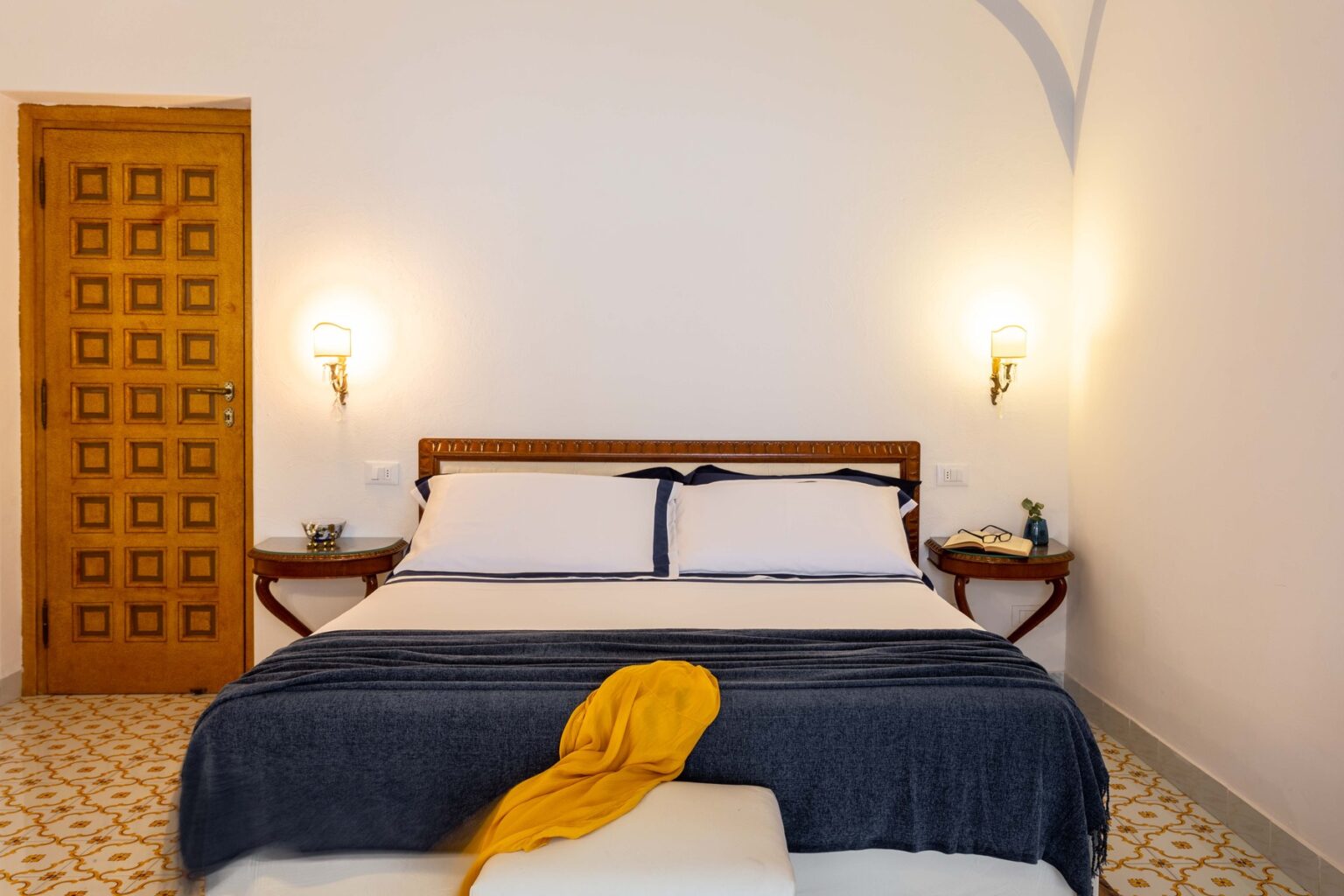 Cozy bedroom featuring a wooden bed with white linens and a navy blue throw, complemented by stylish wall sconces and a decorative door, ideal for a relaxing stay. | Only on SopranoVillas