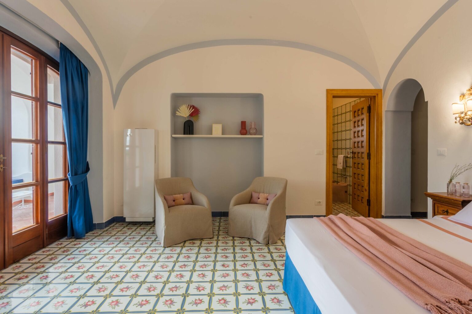 Cozy hotel room featuring two comfortable chairs, a decorative shelf, large windows with blue curtains, and a patterned tiled floor, ideal for relaxation and leisure. | Only on SopranoVillas