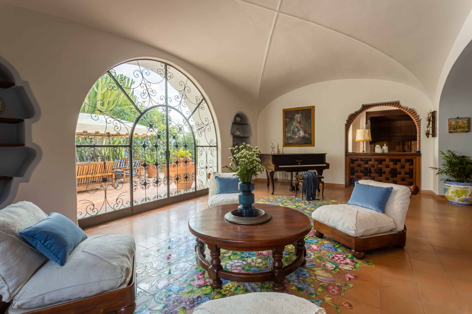 Spacious and inviting living room featuring arched windows, a wooden coffee table, comfortable seating with blue cushions, and a grand piano, surrounded by vibrant floral decor and natural light. | Only on SopranoVillas