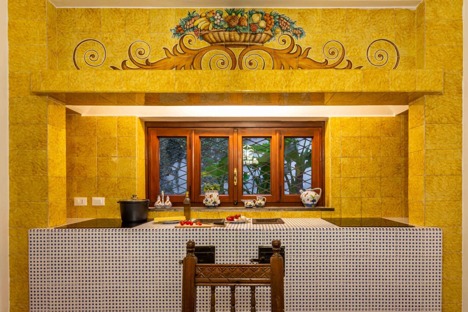 Bright and inviting kitchen featuring yellow tiled walls, a decorative fruit basket mural, and wooden accents. The space includes a traditional wooden chair, a black cooking pot, and elegant ceramic dishware on the counter, creating a warm and welcoming atmosphere. Natural light filters through the window, enhancing the cozy ambiance. | Only on SopranoVillas