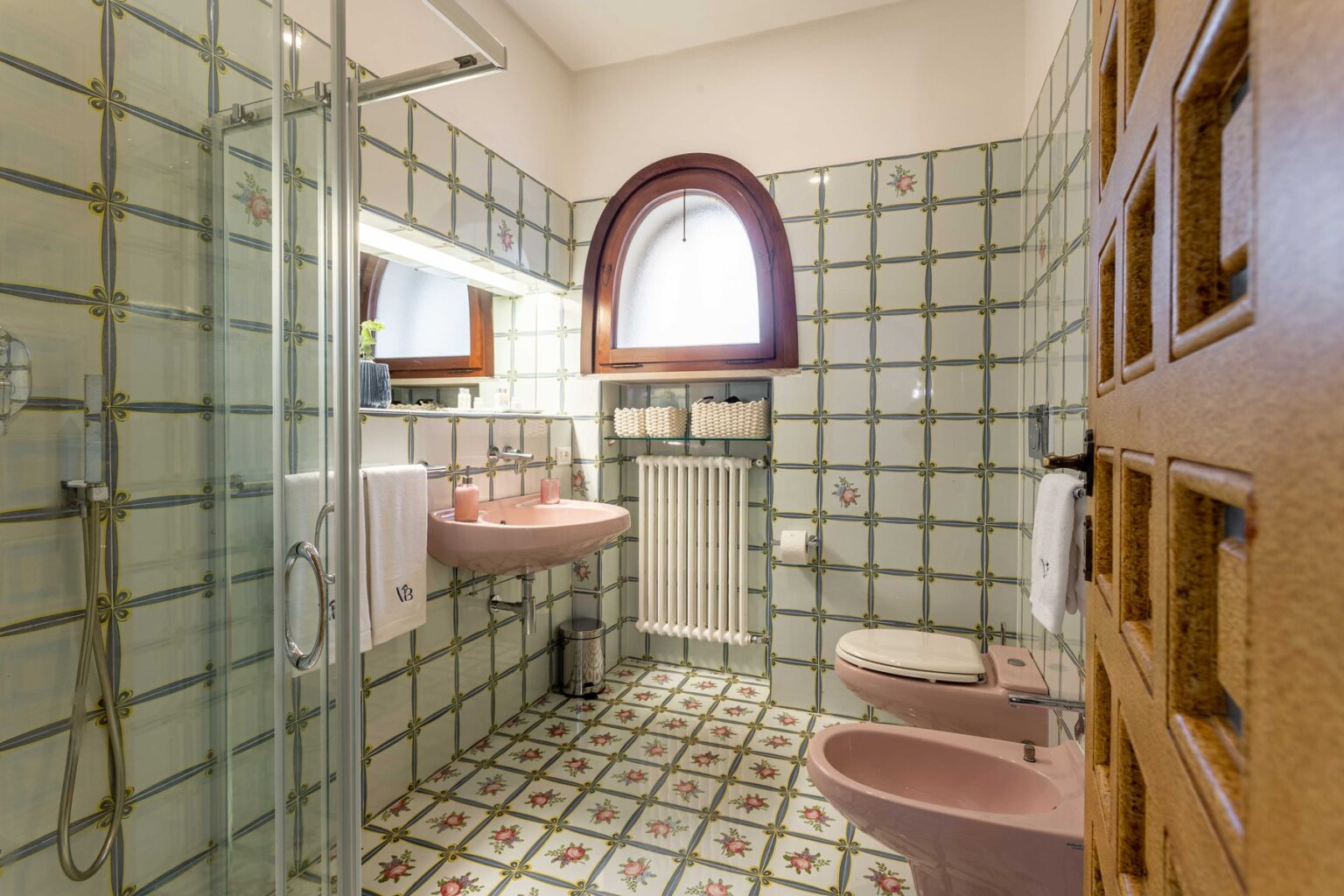 Spacious bathroom featuring vintage-style tiles, a glass shower enclosure, a pink sink, and a wooden door. The decor includes floral patterns and modern amenities, creating a cozy and inviting atmosphere. | Only on SopranoVillas