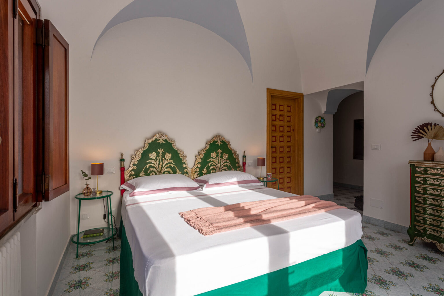 Bright and stylish bedroom featuring a green and gold headboard, white bedding, and a charming vintage dresser. The room is illuminated by natural light from a window, highlighting the decorative floor tiles and cozy furnishings, including a bedside table with a lamp and plant. | Only on SopranoVillas