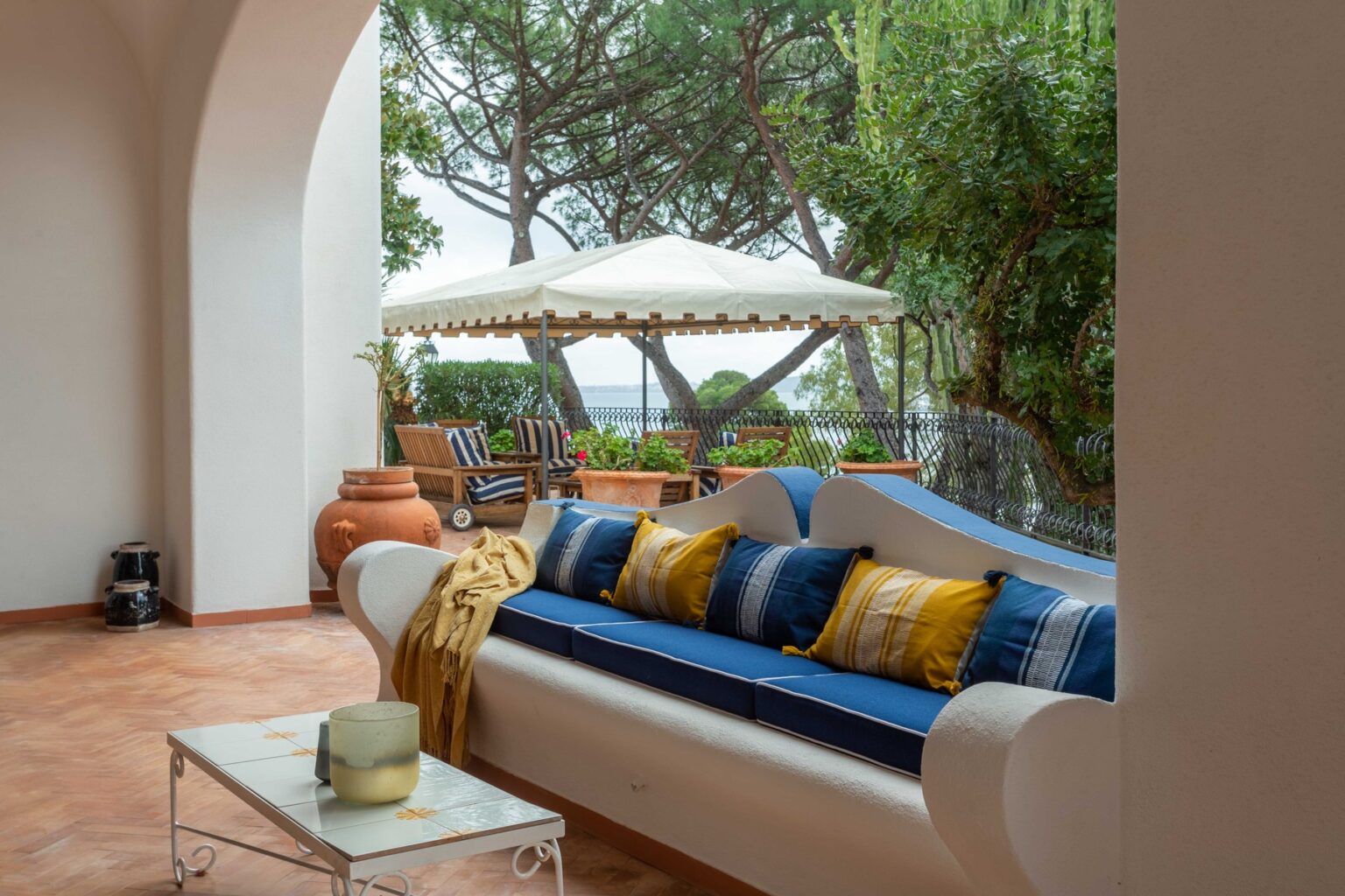 Cozy outdoor seating area featuring a white sofa with blue and yellow cushions, surrounded by greenery and a sunshade, perfect for relaxation. | Only on SopranoVillas