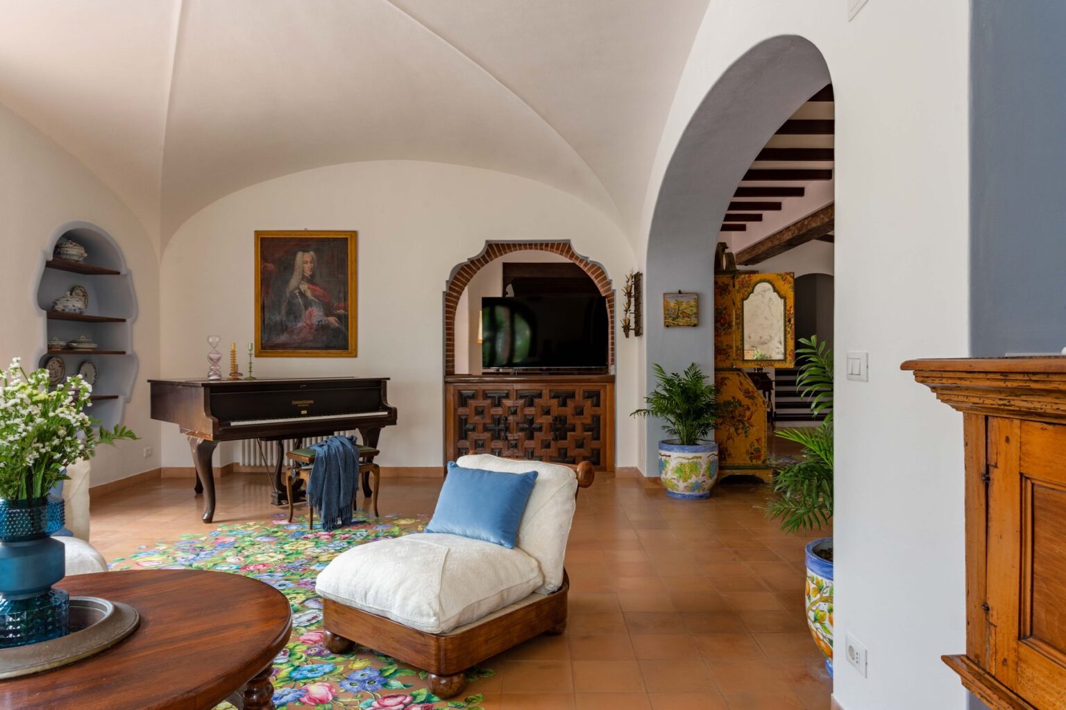 Spacious living room featuring a grand piano, comfortable seating, and decorative plants, with arched doorways and a vibrant area rug, showcasing a blend of elegance and comfort in a well-lit interior. | Only on SopranoVillas