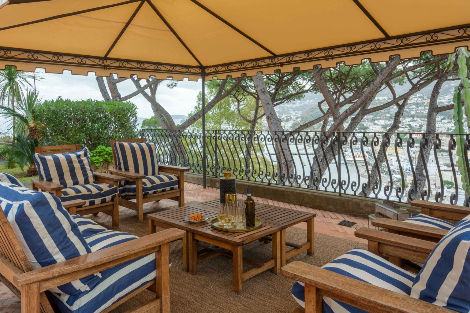 Cozy outdoor seating area under a yellow canopy, featuring striped cushions on wooden chairs, a wooden coffee table with drinks and snacks, and a scenic view of trees and a waterfront cityscape in the background. | Only on SopranoVillas