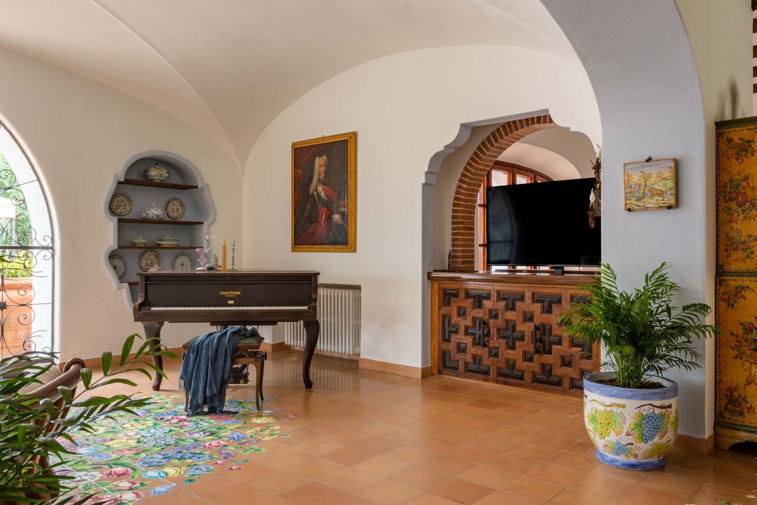 Cozy living room featuring a wooden piano, decorative shelves, and a vintage-style television setup. The space is adorned with plants, colorful area rugs, and artistic wall decor, showcasing an inviting and stylish atmosphere. | Only on SopranoVillas