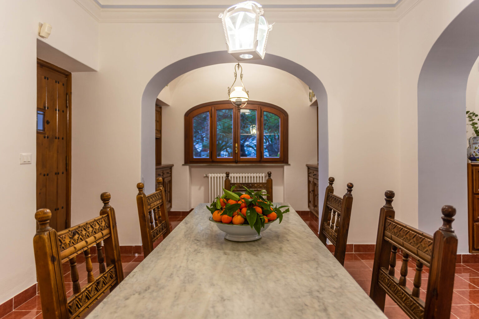 Spacious dining room with a long marble table, wooden chairs, and a bowl of oranges, featuring arched doorways and large windows providing natural light. | Only on SopranoVillas