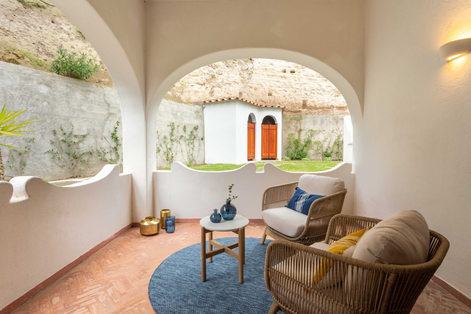 Cozy outdoor seating area featuring two comfortable chairs, a round table with decorative items, and a scenic view of a landscaped garden with an arched stone wall and a rustic building in the background. Ideal for relaxation and enjoying nature. | Only on SopranoVillas