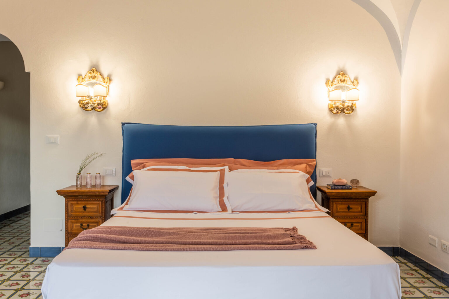 Cozy bedroom featuring a comfortable double bed with a blue headboard, decorative pillows, and elegant bedside tables with lamps, set against a light-colored wall and patterned floor tiles. | Only on SopranoVillas