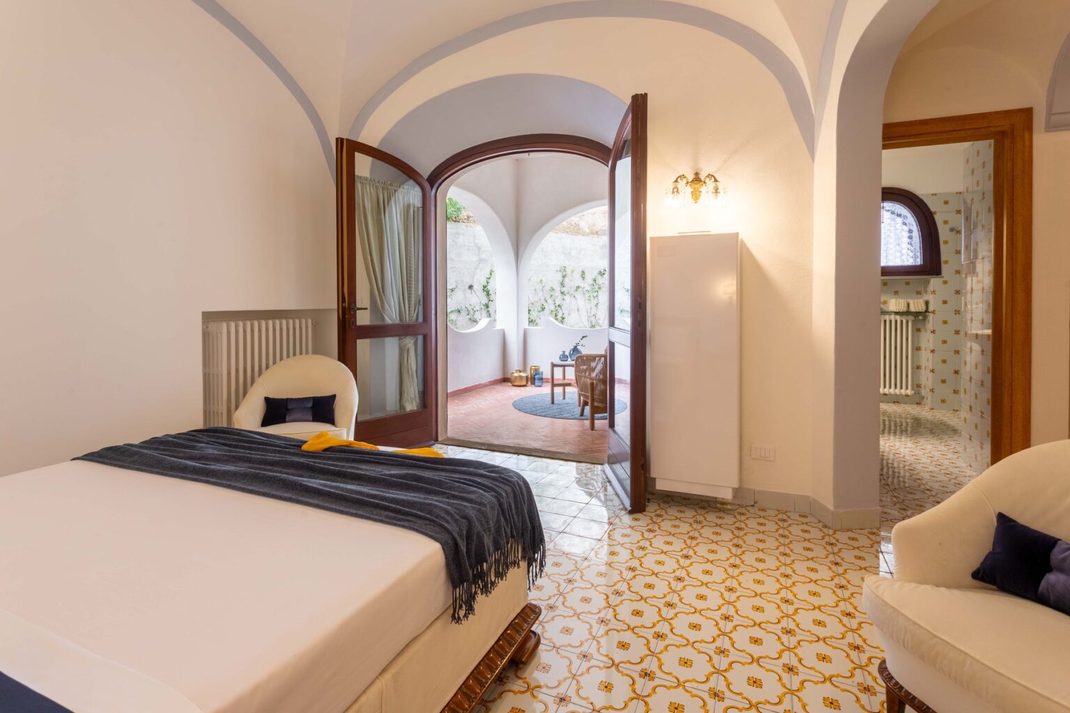 Spacious bedroom with a comfortable bed, elegant decor, and a view of an adjoining living area featuring arched doorways and natural light. The room showcases patterned floor tiles and modern furnishings, creating a cozy atmosphere. | Only on SopranoVillas