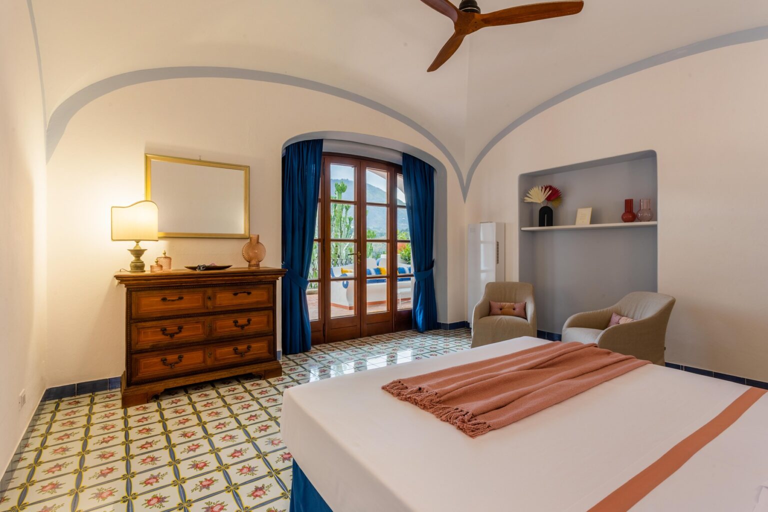 Cozy bedroom with white walls, arched ceiling, and colorful tiled floor. Features a wooden dresser, two armchairs, a lamp, and large windows with blue curtains, providing natural light. | Only on SopranoVillas