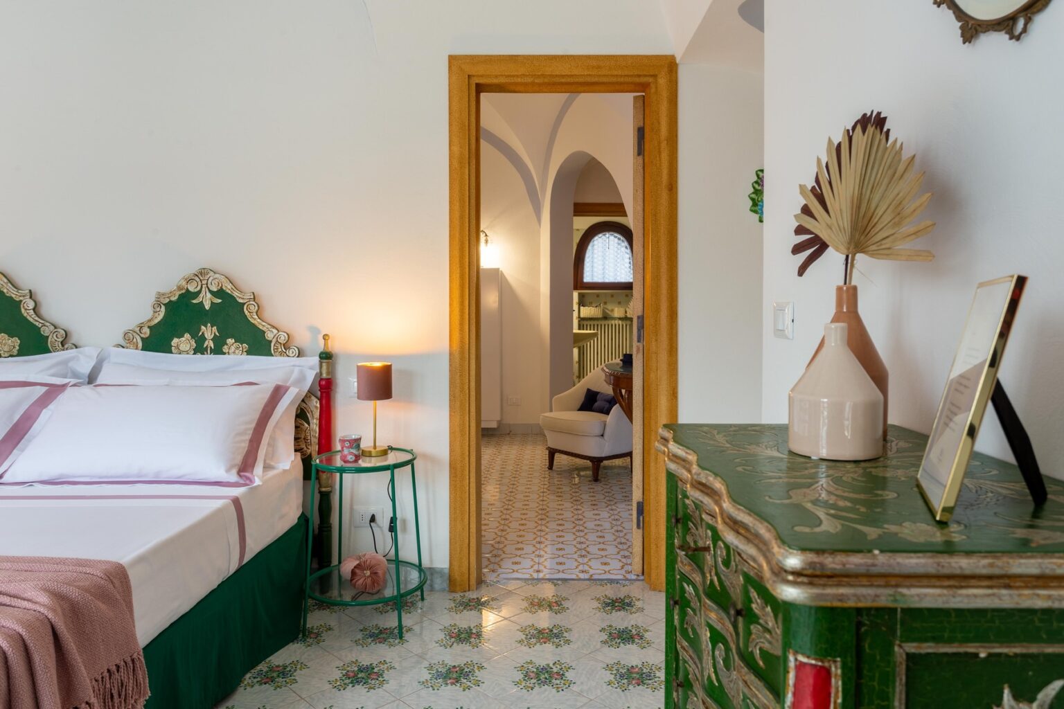 Cozy bedroom interior featuring a green ornate headboard, a bedside table with a lamp, and a decorative dresser. The doorway leads to a bright living area with stylish decor and tiled flooring, highlighting a blend of modern and traditional design elements. | Only on SopranoVillas