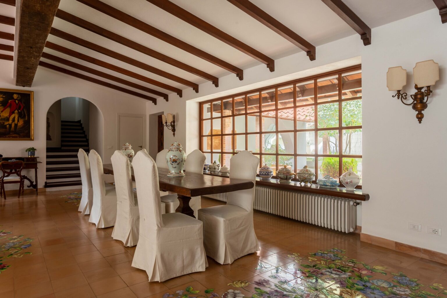 Spacious dining room with a wooden table and white upholstered chairs, featuring exposed wooden beams and large windows that provide natural light, creating a warm and inviting atmosphere. | Only on SopranoVillas