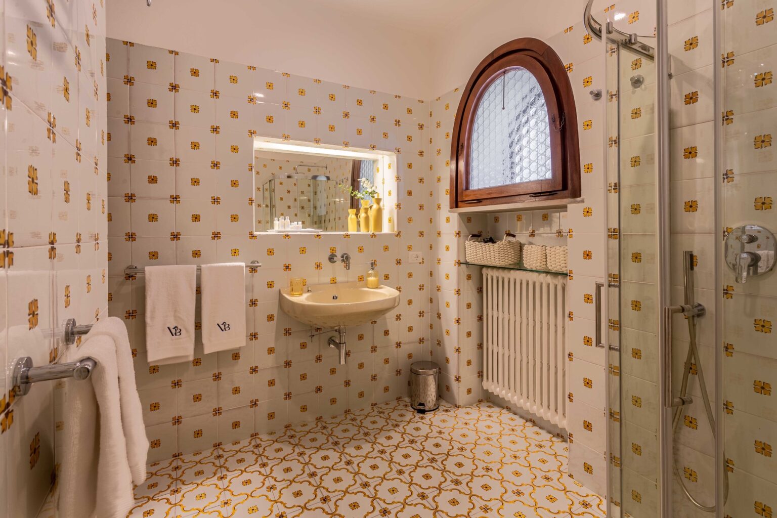 Elegant bathroom featuring patterned tiles, a modern sink, and a glass shower. Natural light enters through an arched window, enhancing the cozy atmosphere. Soft towels are neatly arranged, adding to the inviting decor. | Only on SopranoVillas