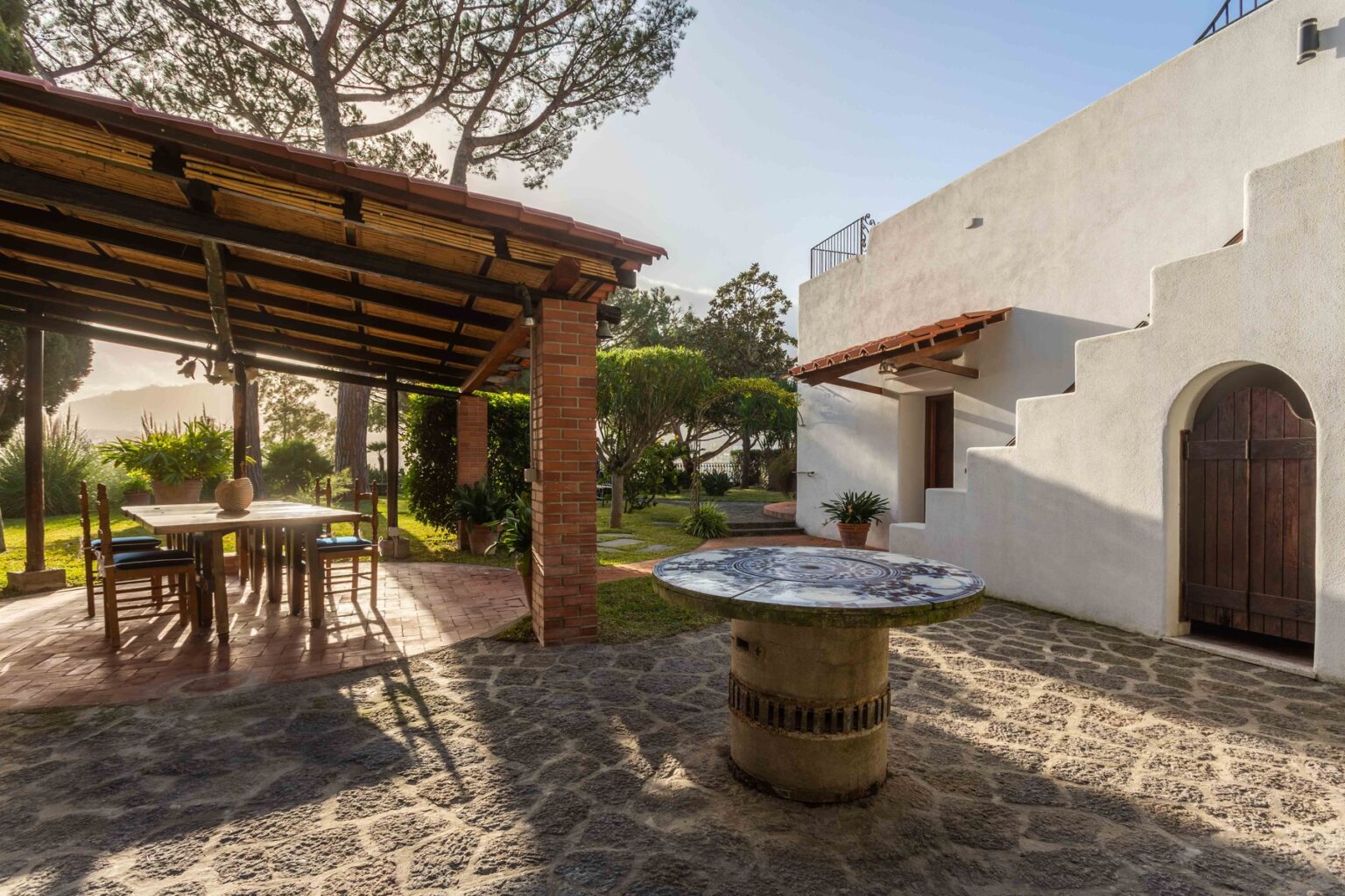 Charming outdoor patio with a rustic dining table and a decorative stone table, surrounded by lush greenery and a modern white house. The scene features a covered area with wooden beams, creating a serene atmosphere perfect for relaxation and outdoor dining. | Only on SopranoVillas