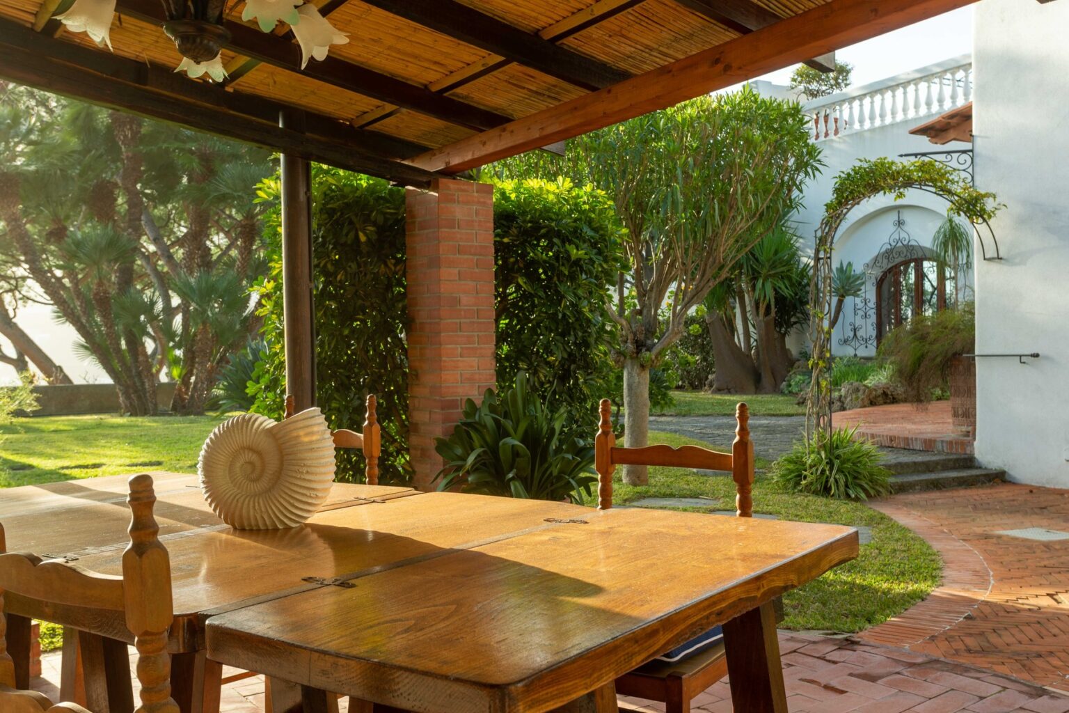 Outdoor dining area with a wooden table and chairs, featuring a decorative shell centerpiece, surrounded by lush greenery and a glimpse of a white building in the background. Ideal for relaxation and enjoying nature. | Only on SopranoVillas
