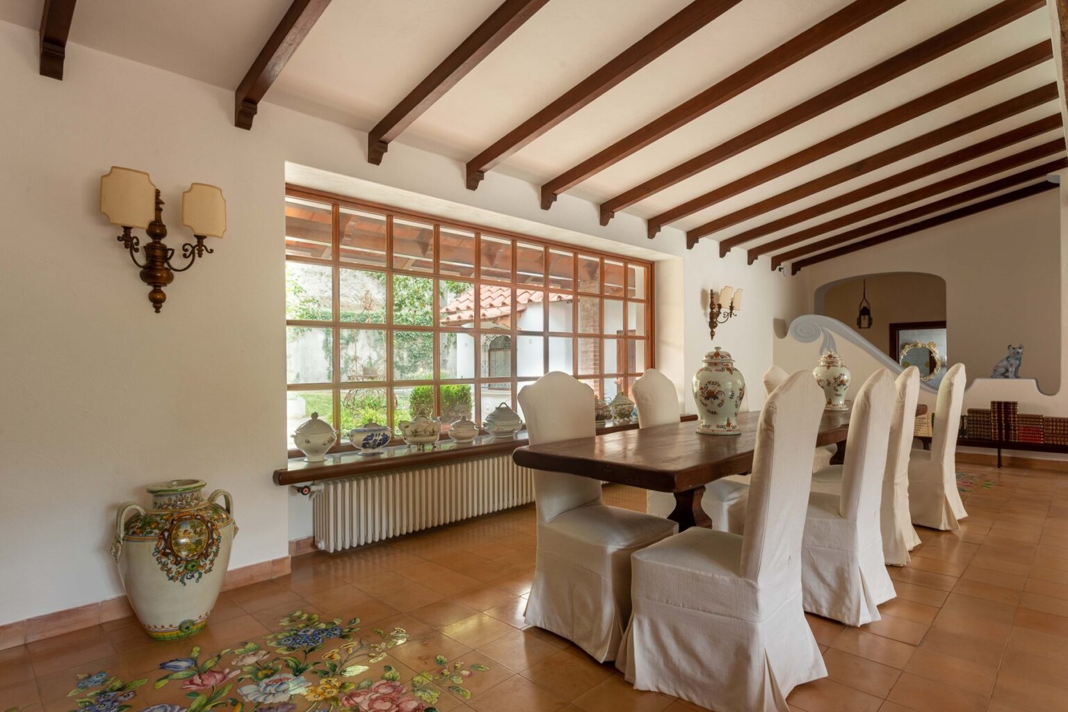 Spacious dining room featuring a long wooden table surrounded by upholstered chairs, large windows allowing natural light, and decorative vases, highlighting a blend of modern and rustic design elements. | Only on SopranoVillas