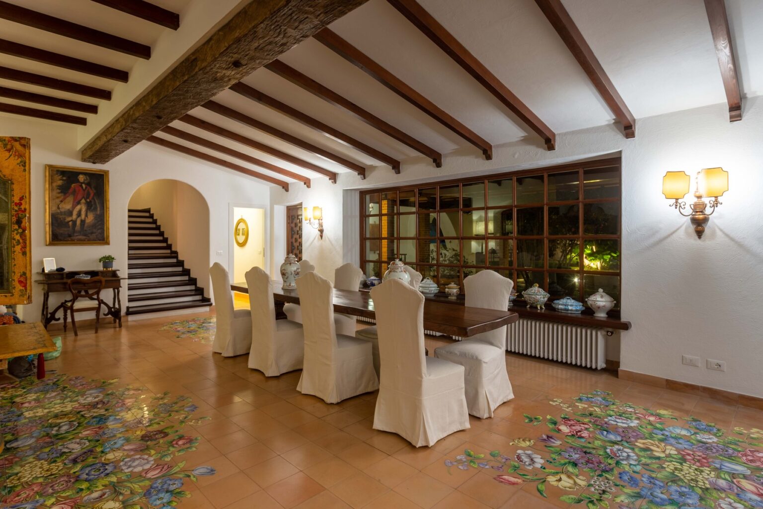 Spacious dining room featuring a long wooden table surrounded by upholstered chairs, rustic wooden beams on the ceiling, and large windows allowing natural light. Colorful tiled floor adds a vibrant touch to the elegant decor. | Only on SopranoVillas
