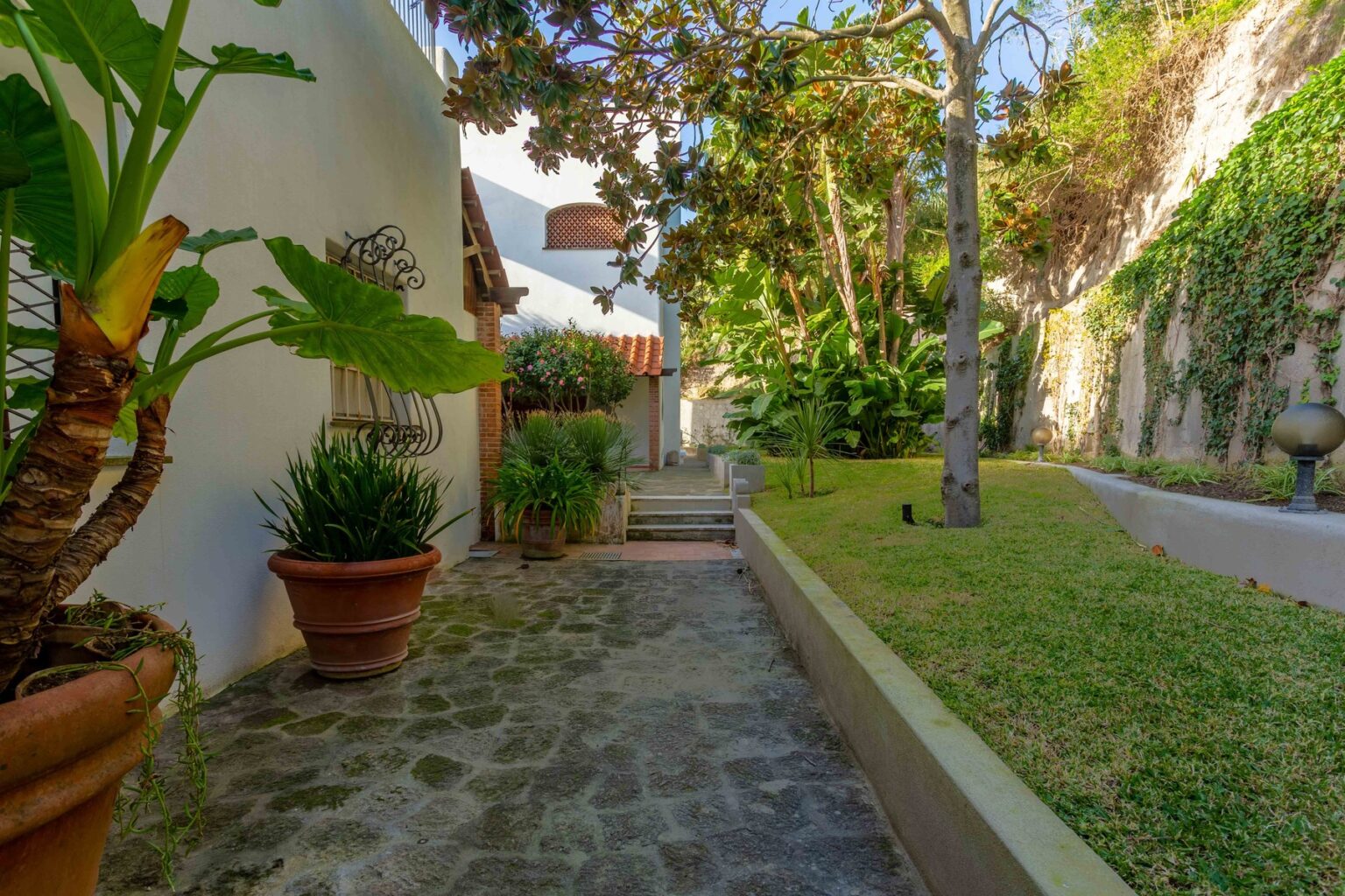 Lush garden pathway with stone paving, bordered by potted plants and greenery, leading to a serene outdoor space. Ideal for showcasing landscaping design and outdoor living areas. | Only on SopranoVillas
