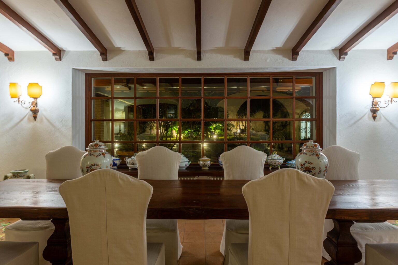 Elegant dining room featuring a long wooden table surrounded by white upholstered chairs, with a large window showcasing lush greenery outside. Soft lighting from wall sconces enhances the warm ambiance. | Only on SopranoVillas