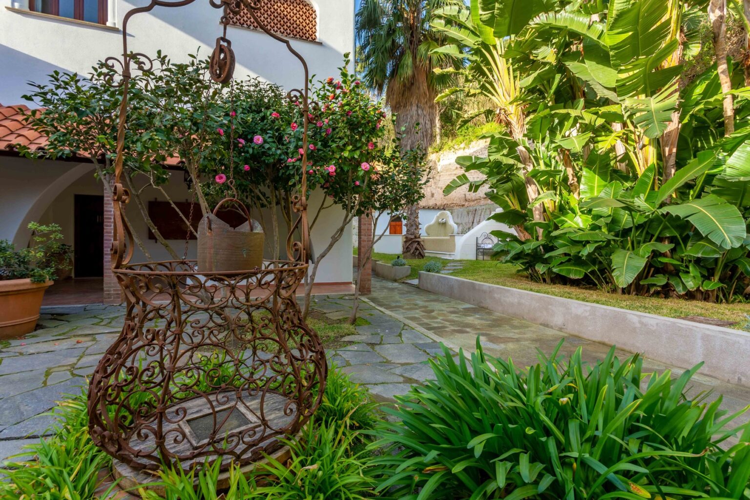 Rustic outdoor garden featuring a decorative hanging chair surrounded by lush greenery and tropical plants, with a stone pathway leading to a charming building. Ideal for relaxation and outdoor living. | Only on SopranoVillas