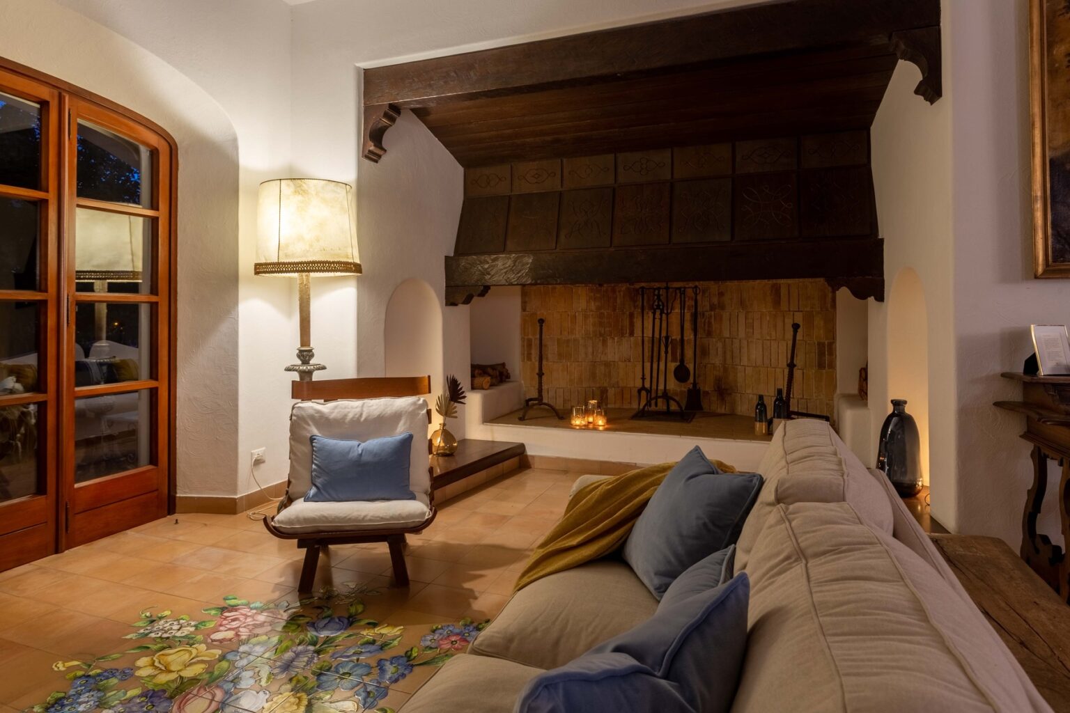 Cozy living room interior featuring a beige sofa with colorful pillows, a wooden beam ceiling, a rustic fireplace, and a stylish floor lamp, creating a warm and inviting atmosphere. | Only on SopranoVillas