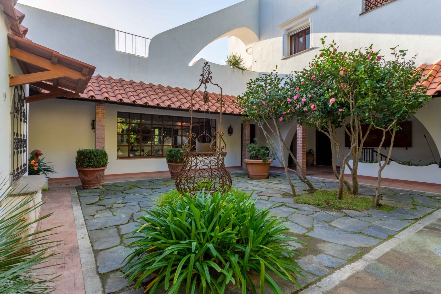 Courtyard with stone flooring, lush greenery, and potted plants, featuring a traditional Mexican tile roof and an inviting, tranquil atmosphere. | Only on SopranoVillas