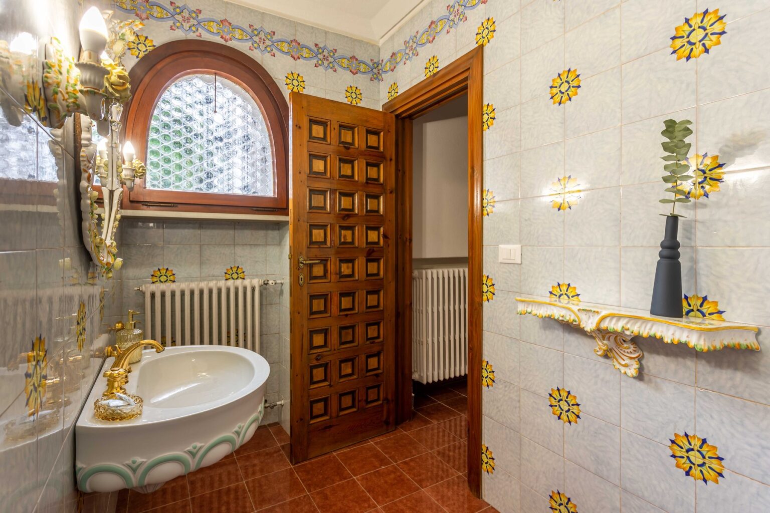 Elegant bathroom featuring vintage decor with ornate golden fixtures, a stylish basin, and colorful tile work. The room includes a wooden door, a decorative shelf, and a classic radiator, all complemented by a beautiful window design that allows natural light to enter. | Only on SopranoVillas