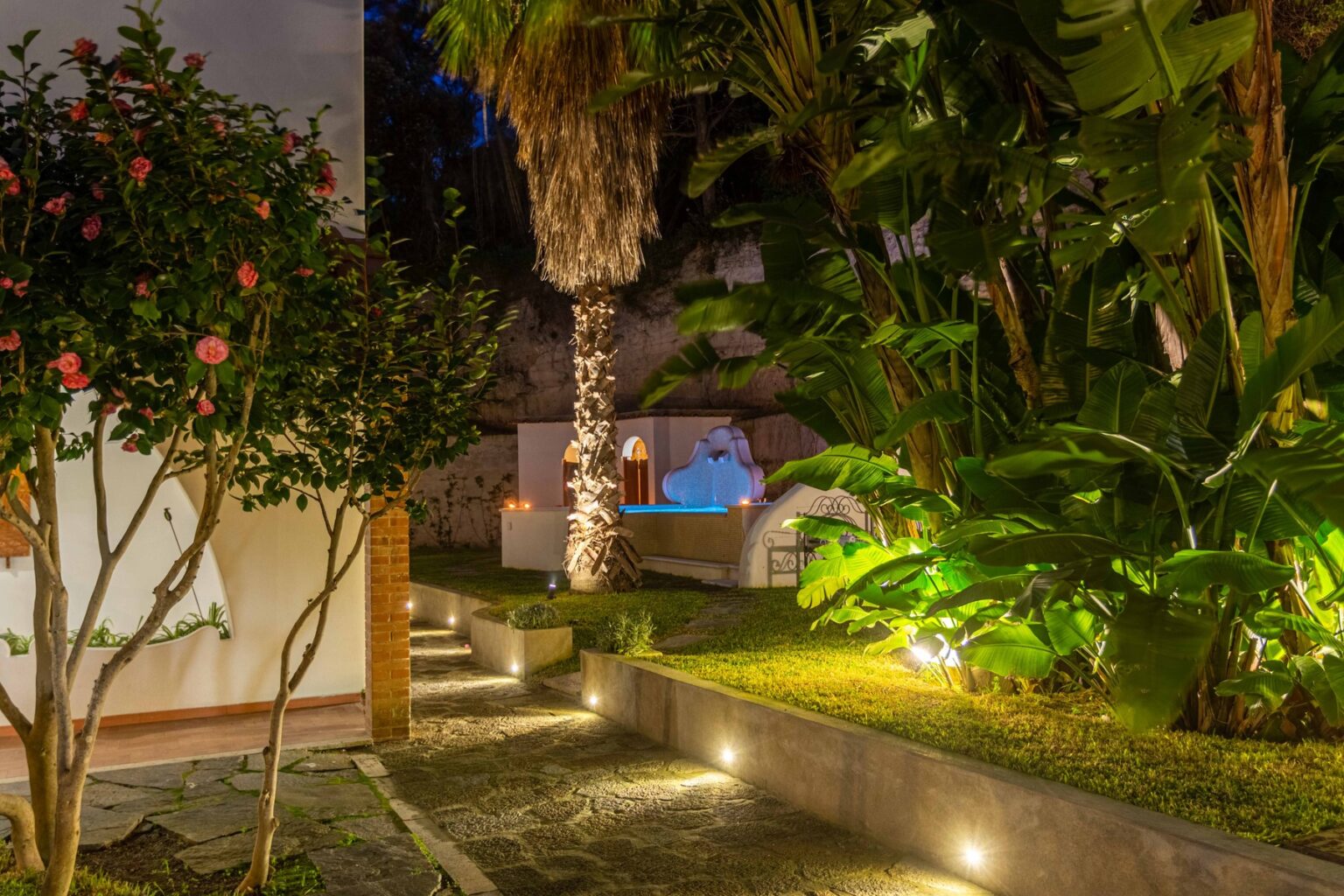 Lush garden at night featuring vibrant flowers, palm trees, and a softly illuminated pathway leading to a serene pool area surrounded by tropical plants. | Only on SopranoVillas