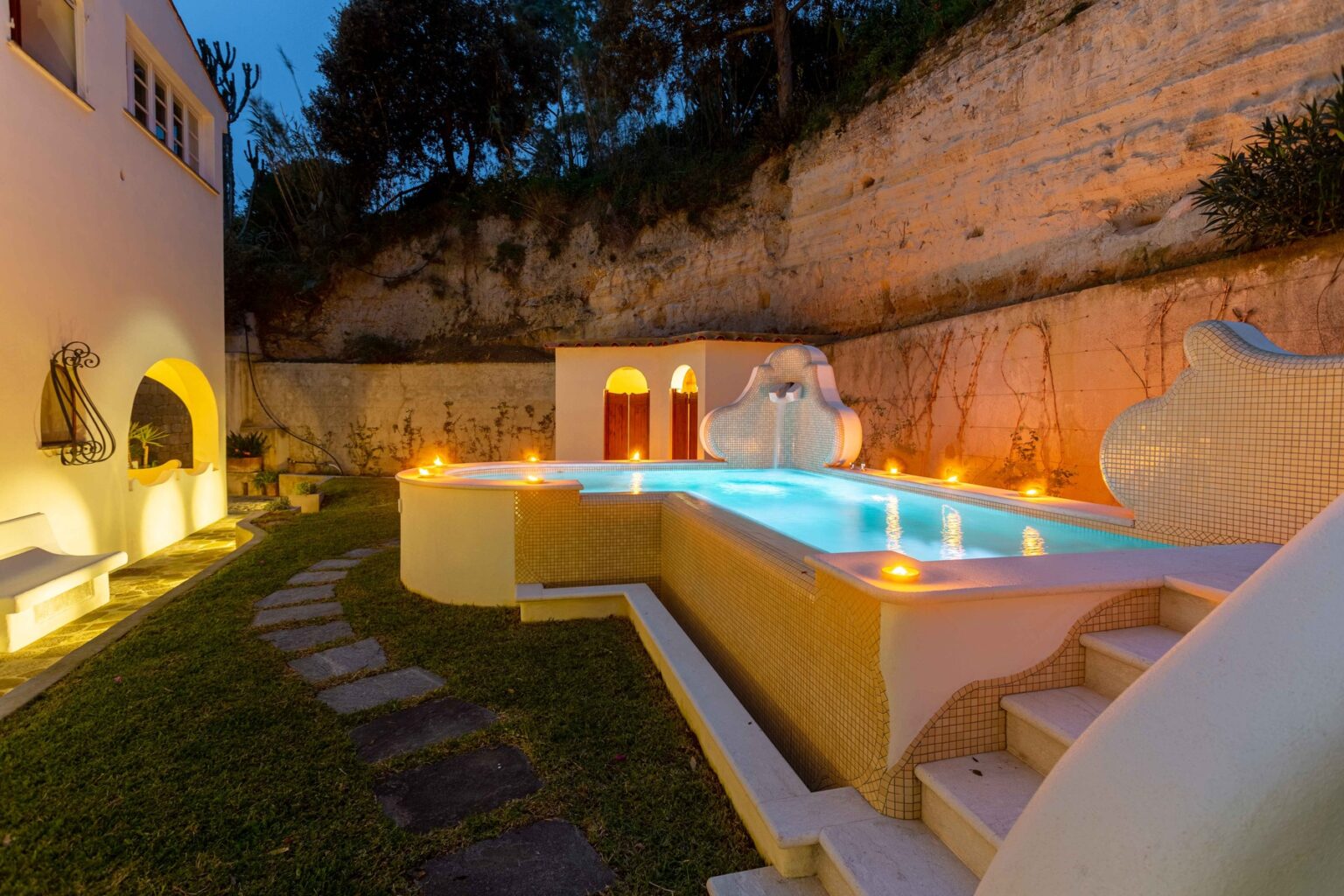 Serene outdoor pool area at dusk, featuring illuminated pathways, a modern design with mosaic tiles, and surrounding greenery, perfect for relaxation and entertainment. | Only on SopranoVillas