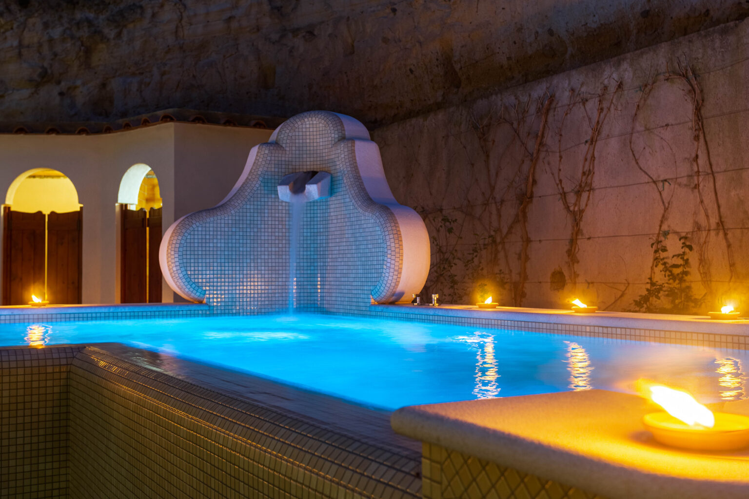 Luxurious indoor pool with a decorative tiled fountain, illuminated by soft candlelight, surrounded by stone walls and arches, creating a serene and inviting atmosphere for relaxation. | Only on SopranoVillas