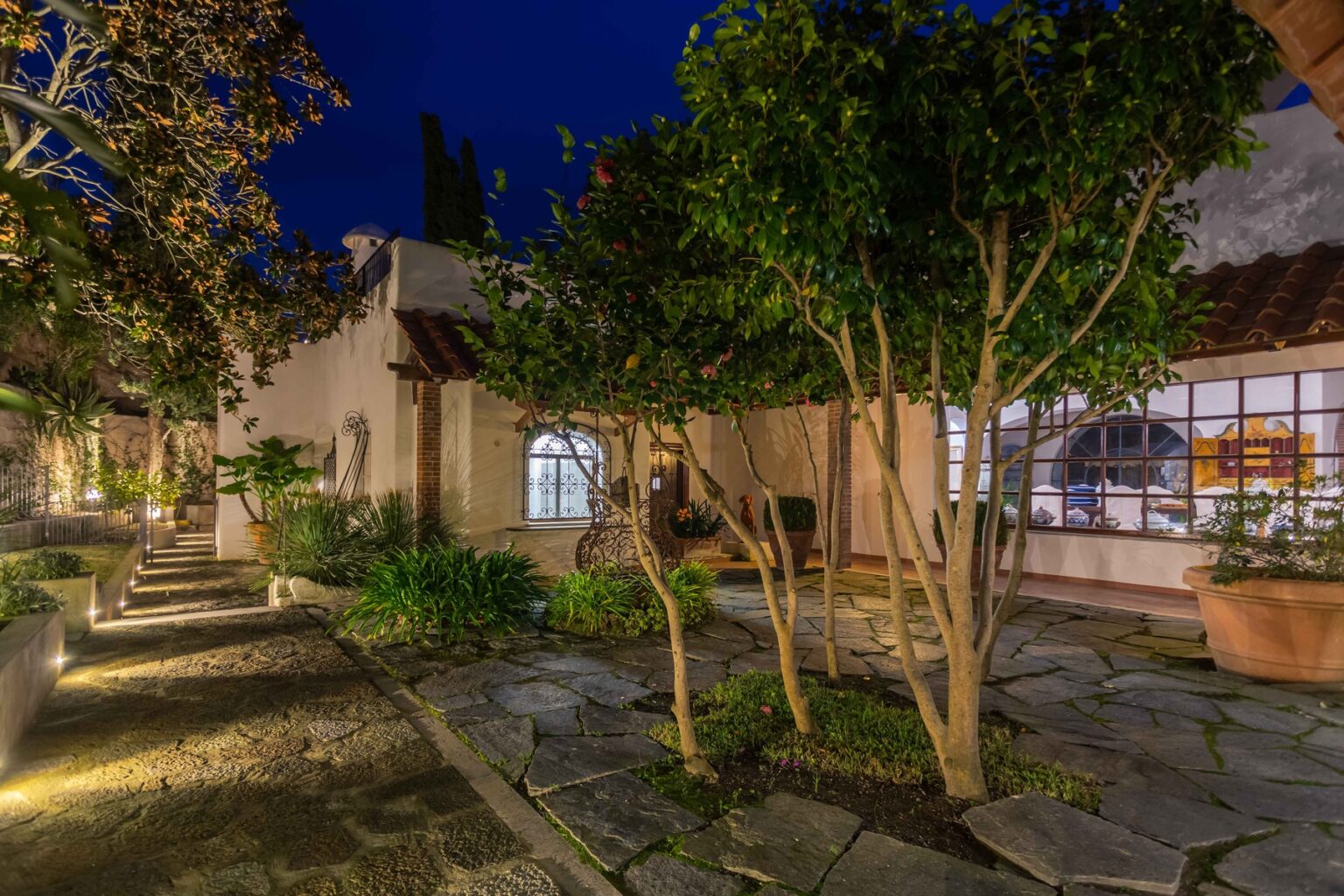 Lush garden pathway with stone tiles leading to a beautifully lit home, featuring green trees and plants, under a night sky. The architecture showcases a blend of rustic and modern design elements. | Only on SopranoVillas