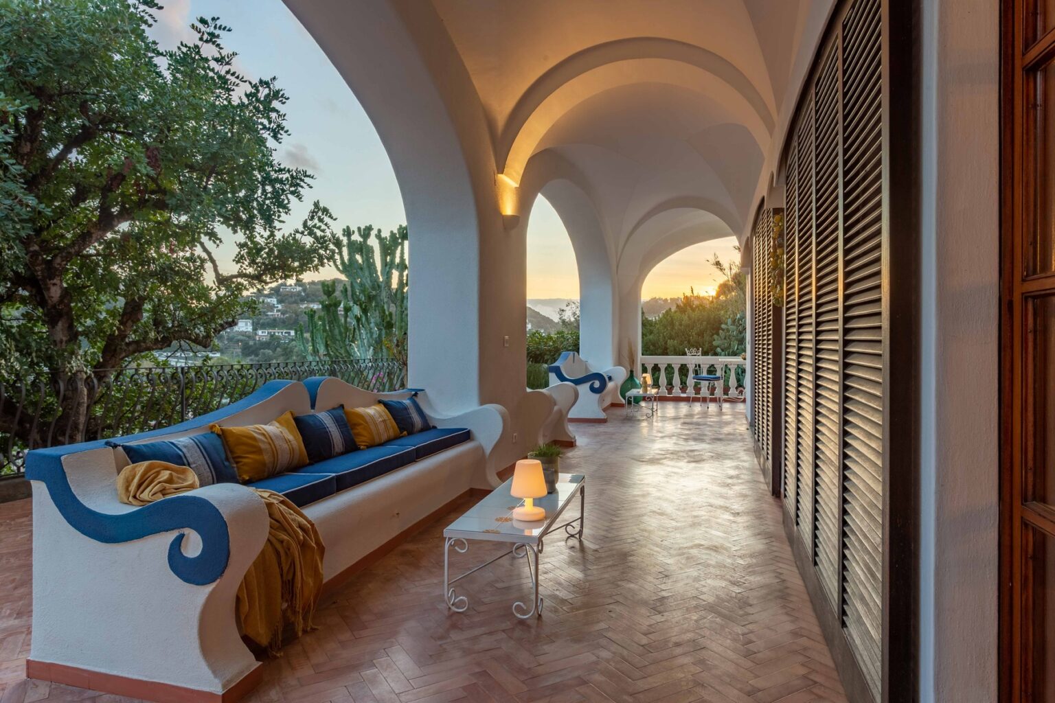 Elegant outdoor terrace with a comfortable sofa adorned with colorful cushions, featuring arched architecture and lush greenery, illuminated by warm evening light. Perfect for relaxation and enjoying sunset views. | Only on SopranoVillas