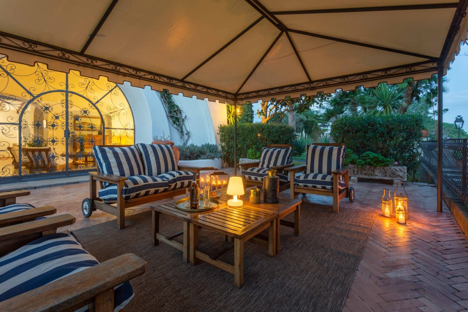 Cozy outdoor seating area under a tent, featuring striped lounge chairs, a wooden coffee table, and ambient lanterns, surrounded by greenery during twilight. | Only on SopranoVillas