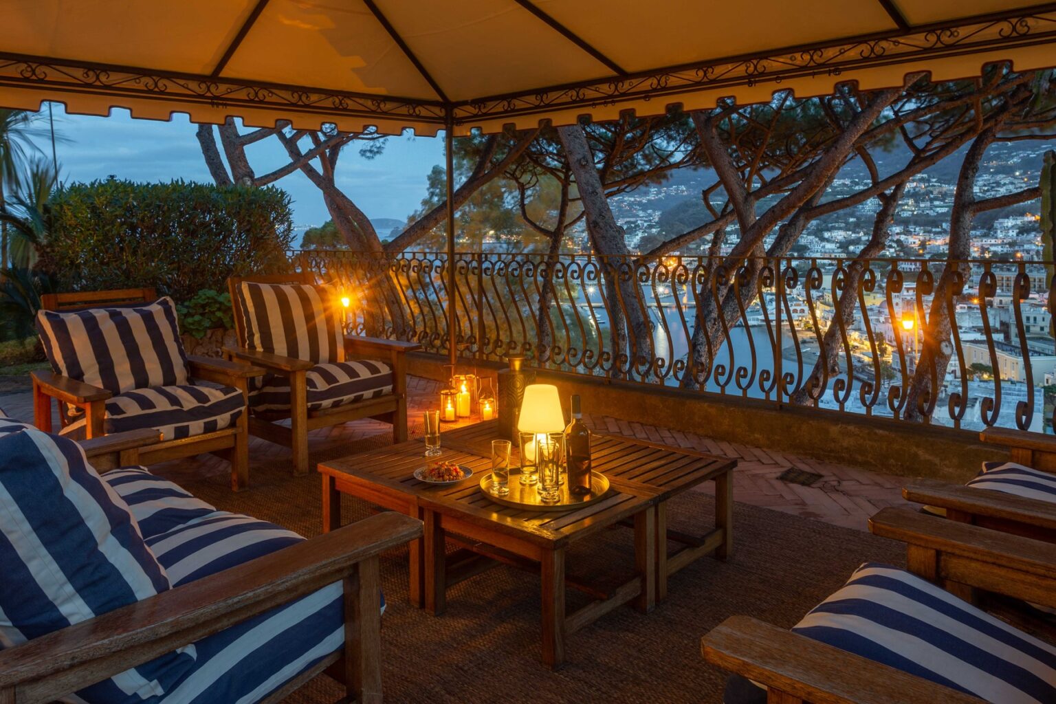 Cozy outdoor seating area with striped chairs and a wooden table, illuminated by soft candlelight, overlooking a scenic view at dusk. | Only on SopranoVillas