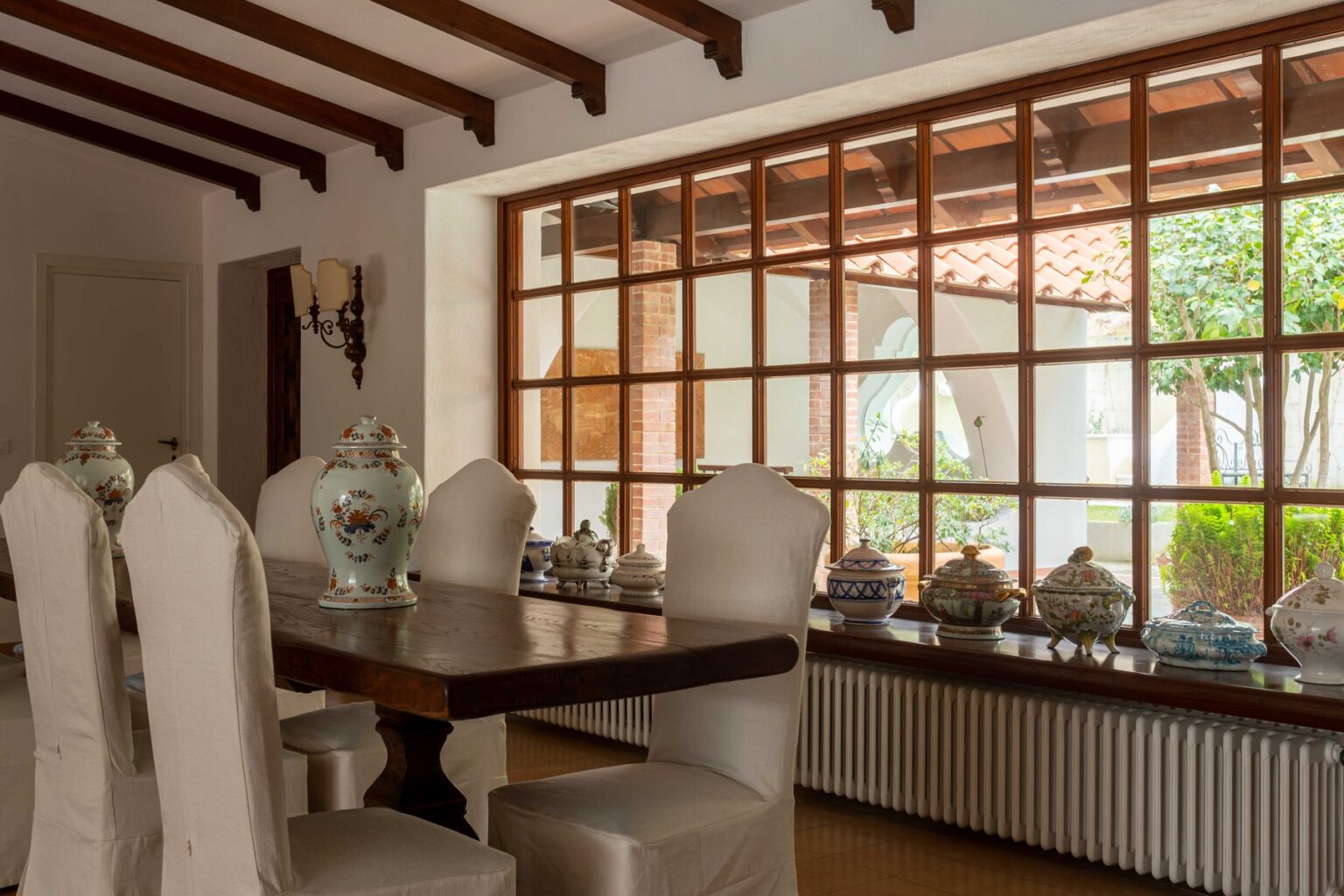 Elegant dining room featuring a wooden table and upholstered chairs, complemented by a large window with wooden frames that offers a view of a garden. Decorative porcelain items are displayed on the windowsill, enhancing the sophisticated ambiance. | Only on SopranoVillas