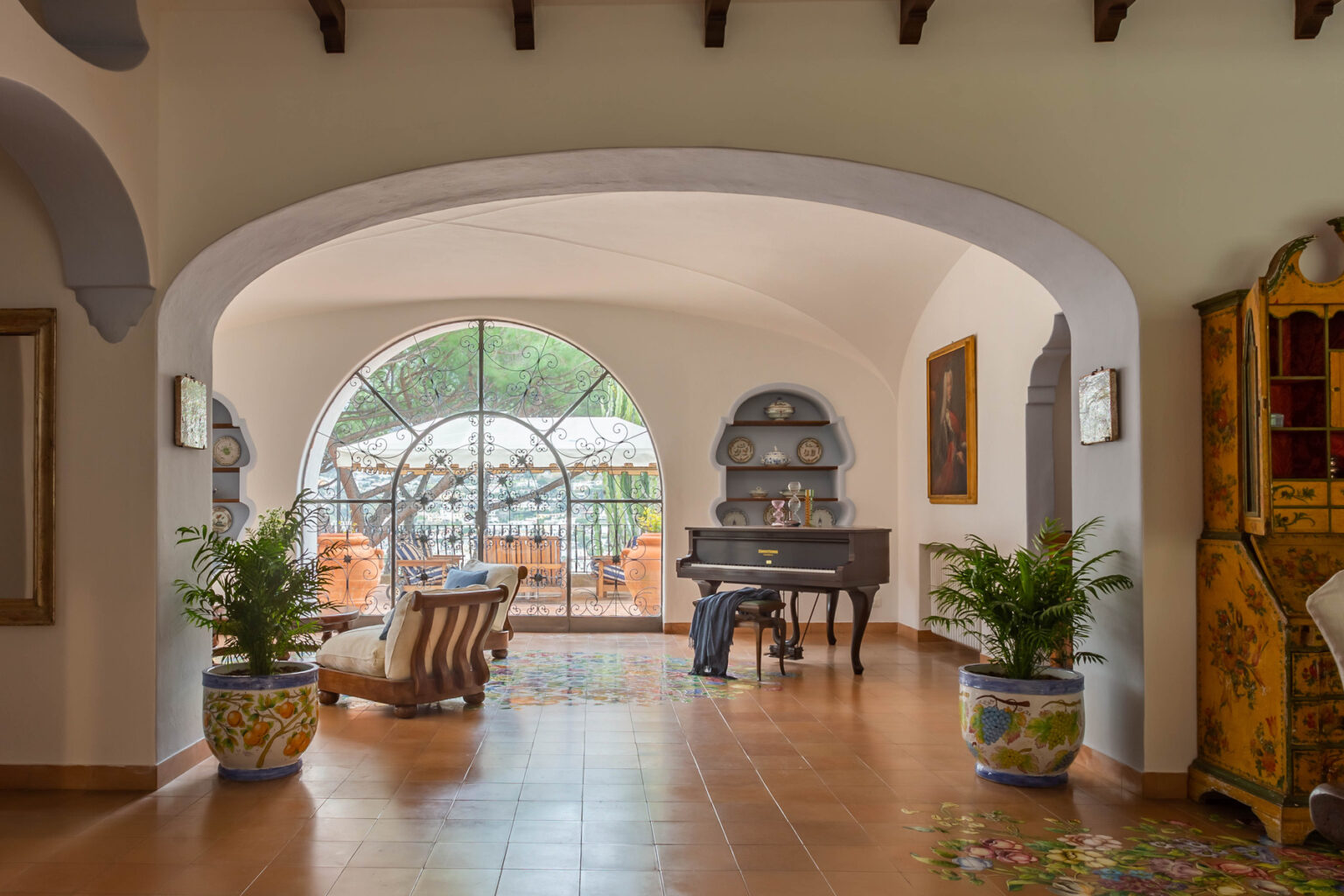 Elegant interior of a spacious living room featuring a grand piano, comfortable seating, and large arched windows that open to a terrace. Decorative plants and antique furniture add warmth and charm to the inviting space. | Only on SopranoVillas