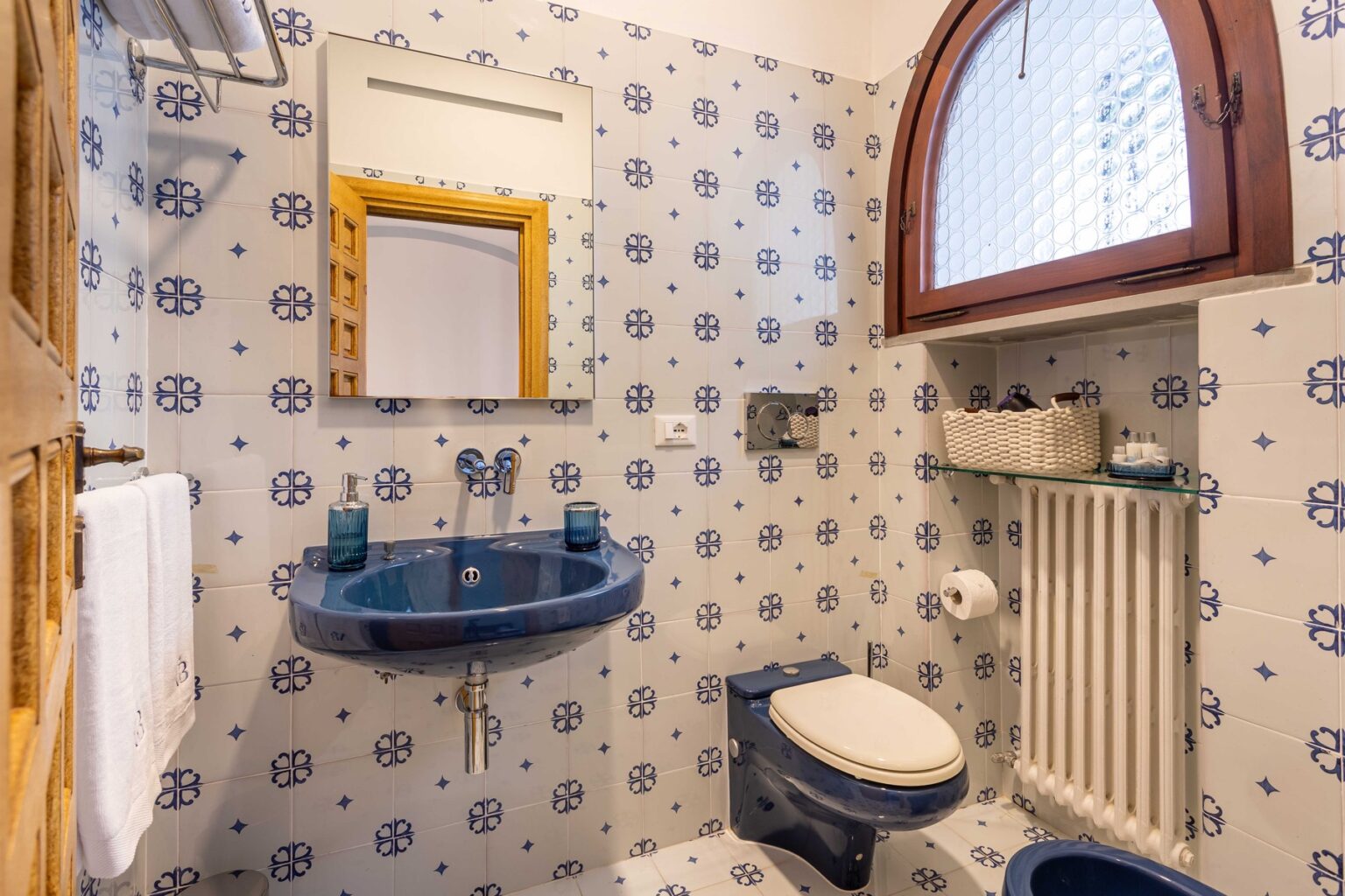 Charming bathroom with blue patterned tiles, a round mirror, a blue sink, and a white toilet, featuring natural light from an arched window. | Only on SopranoVillas