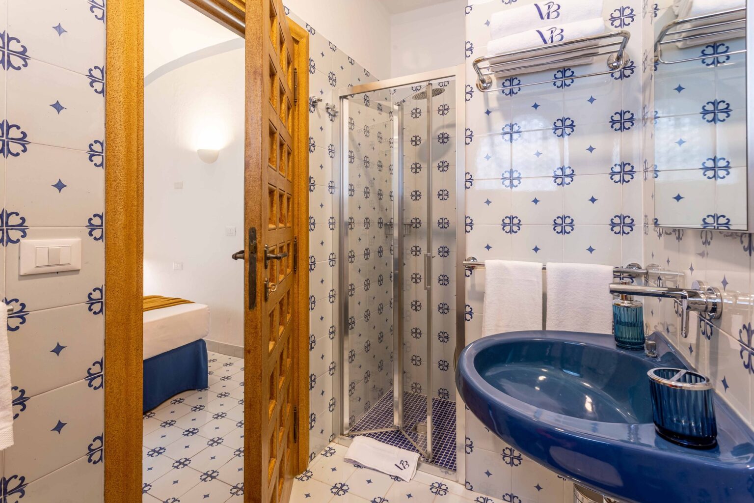 Modern bathroom featuring a blue sink, glass shower, and decorative tilework with blue and white patterns. The wooden door adds a rustic touch, while plush towels are neatly arranged on a rack. | Only on SopranoVillas