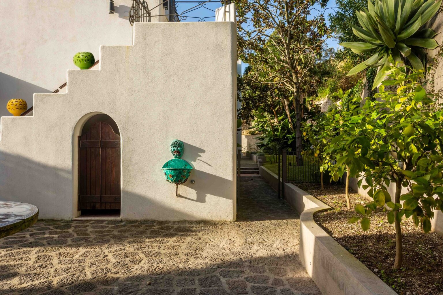 Colorful decorative pots line the staircase of a modern outdoor courtyard, featuring a rustic wooden door and a unique turquoise water fountain. Lush greenery and stone pathways enhance the vibrant aesthetic of the space. | Only on SopranoVillas