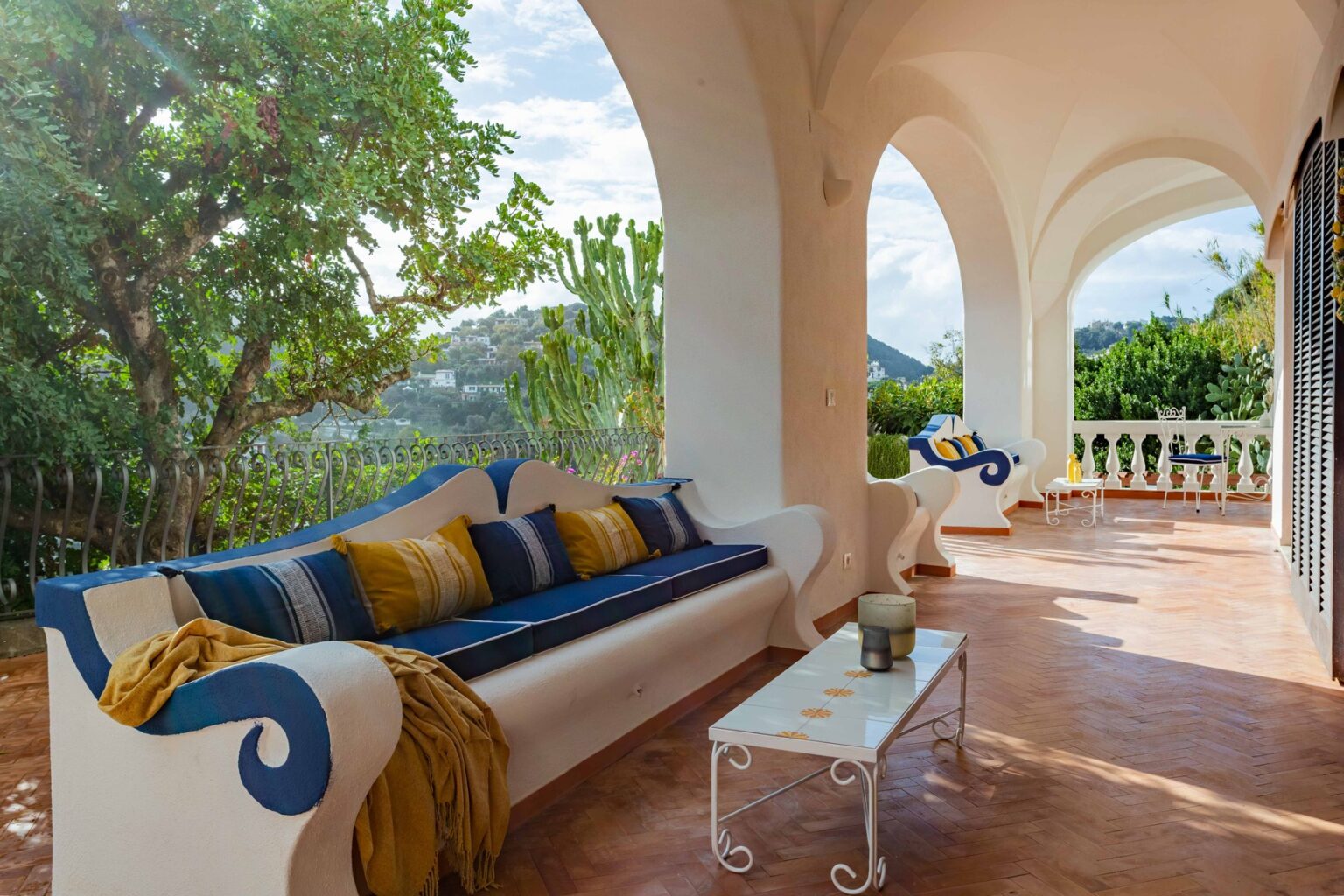 Cozy outdoor terrace featuring a stylish white sofa with blue and yellow cushions, a small coffee table, and lush greenery in the background, perfect for relaxation and enjoying scenic views. | Only on SopranoVillas