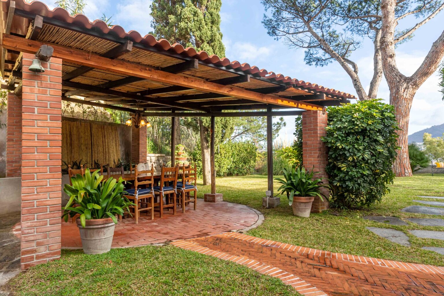 Outdoor dining area with a rustic brick pergola, surrounded by lush greenery and potted plants, offering a serene atmosphere for gatherings. | Only on SopranoVillas