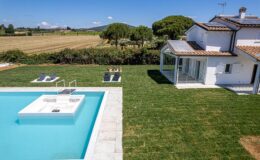 Modern villa with a swimming pool and sun loungers in a lush green garden, surrounded by scenic countryside and clear blue skies. Ideal for relaxation and outdoor living.