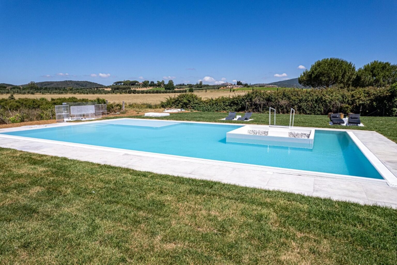 Modern outdoor swimming pool surrounded by lush green grass and scenic countryside, featuring a spacious deck and lounge chairs under a clear blue sky. | Only on SopranoVillas