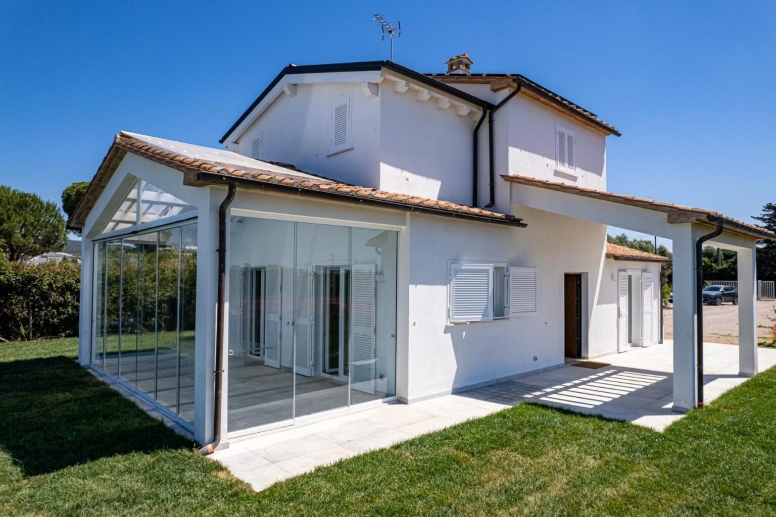 Modern white house with a glass extension, surrounded by green grass and clear blue sky, showcasing contemporary architectural design. | Only on SopranoVillas